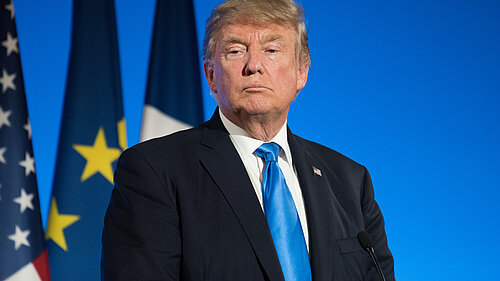 An image of Donald Trump standing in front of a blue backdrop with the US, EU, and French flags behind him
