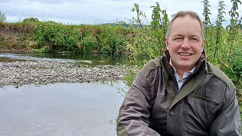 A photo of Richard Foord crouched down by a river. Behind him is a small island made of stones and some greenery.