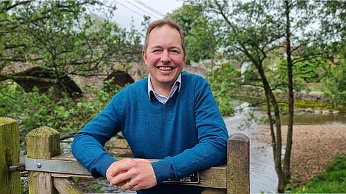 Richard Foord standing leaning on a wooden gate. He is facing the camera and smiling. He is wearing a blue jumper. Behind him are an old stone bridge, a stream, and some trees.