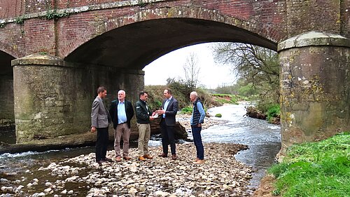 A photo of a group of people standing on a rocky exposed section of riverbed by an old stone bridge.