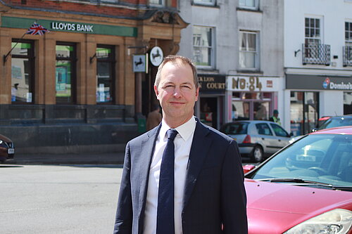 A photo of Richard Foord wearing a dark suit and standing across the road from Lloyds bank in Honiton on a bright day.