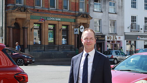 A photo of Richard Foord wearing a dark suit and standing across the road from Lloyds bank in Honiton on a bright day.