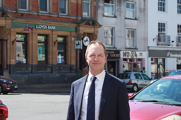 A photo of Richard Foord wearing a dark suit and standing across the road from Lloyds bank in Honiton on a bright day.