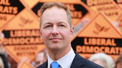 Head and shoulders image of Richard Foord in front of activists holding signs reading "Liberal Democrats winning here"