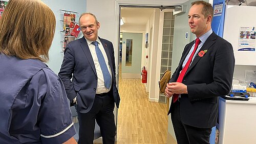 Ed Davey and Richard Foord standing in the corridor of a medical building, speaking to a nurse who has her back to the camera. Ed Davey is on the left in a suit with a bue tie, and Richard on the right in a suit with a red tie and a Remembrance poppy