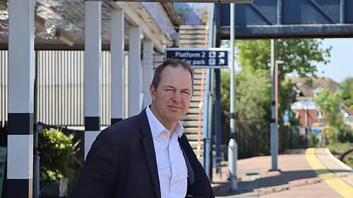 A photo of Richard Foord MP standing on the platform in front of the ticket office at Honiton Station on a sunny day