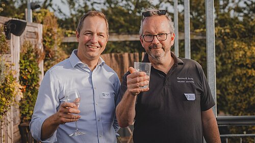 A photo of two men taken in a vineyard on a summer evening. On the left is Richard Foord and the right Ruud Jansen Venneboer. Both men are looking toward the camera and holding up glasses.