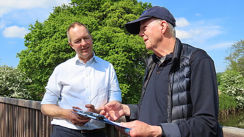A photo of Richard Foord MP (left) speaking to a representative of the Otter Valley Association. Both men are stood on a wooden bridge with their backs to the railing. There is a tree behind them and the weather is clear and sunny.