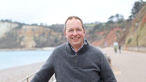 A photo of Richard Foord on the seafront at Seaton. The background is out of focus, with the rest of the promenade and cliffs in the background. There are also a couple of people visible in the distance. The weather appears overcast.