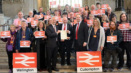 A group of people standing on the steps in Westminster Hall holding signs with the National Rail logo. There are two larger signs on the floor with the logo and with the names "Wellington" and "Cullompton" respectively. In the centre are Gideon Amos MP, Rail Minister Lord Hendy, and Richard Foord MP.