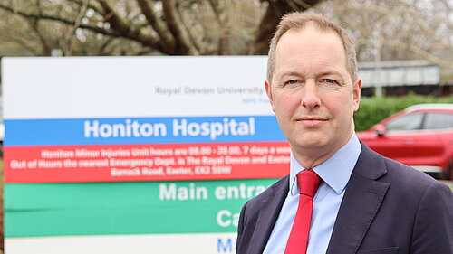 A photo of Richard Foord wearing a suit and standing in front of the sign for Honiton Hospital, which is out of focus